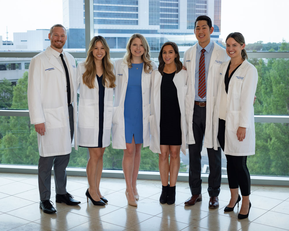 To men and 4 women wearing lab coats over dress clothes, standing in front of a large window with a view of a high-rise building.